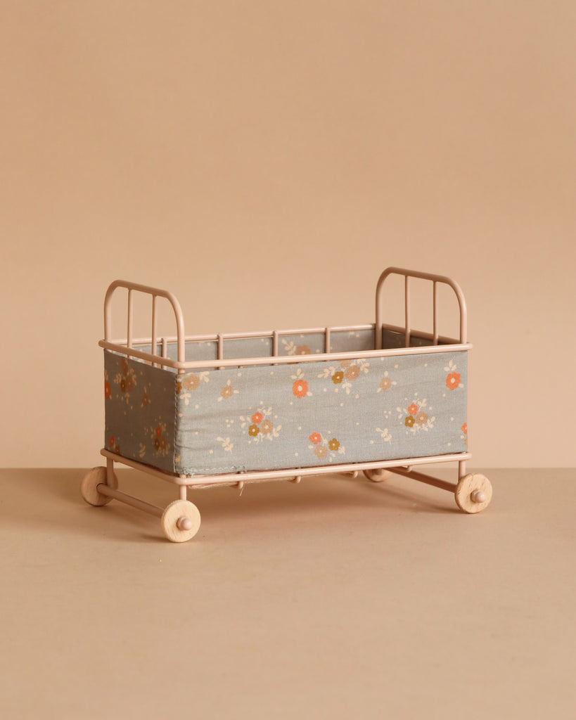 The Maileg Cot Bed, Micro - Powder features a light pink metal frame with wheels and blue floral fabric sides, and is shown on a beige surface against a matching beige background.