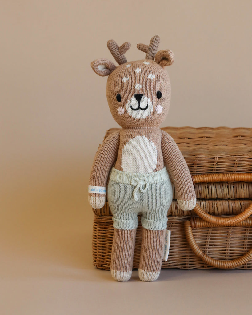 Cuddle + Kind Elliott the Fawn, a hand-knit deer plush with antlers and light blue shorts, rests against a wicker basket on a beige background—crafted as a sustainable, fair trade treasure.