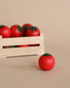 A wooden crate filled with bright red Erzi Tomato Pretend Food tomatoes stands on a soft beige background, with one tomato placed in front of the crate.