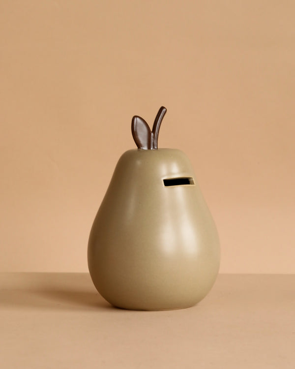 The Ferm Living Pear Money Bank is a glazed ceramic pear-shaped piggy bank with a top coin slot and two small brown leaves—ideal for kids’ room decor, pictured on a beige background.