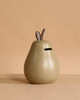 The Ferm Living Pear Money Bank is a glazed ceramic pear-shaped piggy bank with a top coin slot and two small brown leaves—ideal for kids’ room decor, pictured on a beige background.