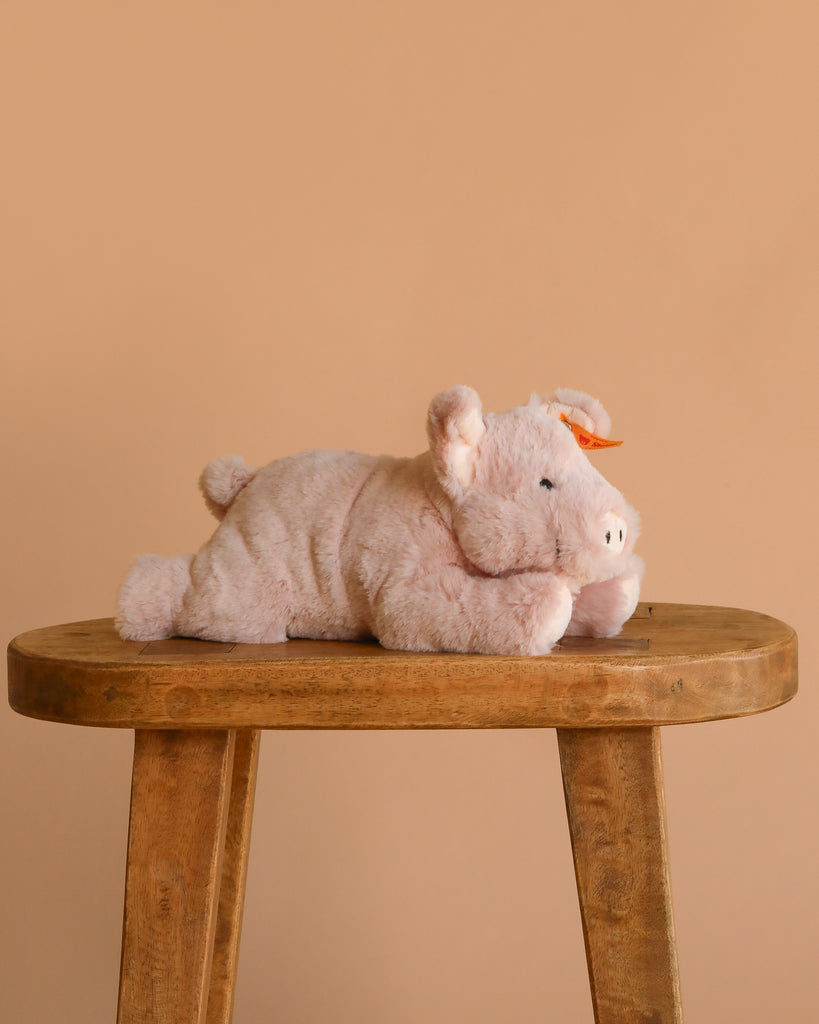 The Steiff Piko Pig Plush Toy, 11 inches, features soft light pink fur and a small orange bow on its ear. It rests on a wooden stool against a beige background—an adorable addition to any stuffed animal collection.