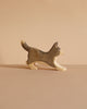 The Handmade Wooden Border Collie is a small, handcrafted dog figurine with a raised tail, painted in brown and beige plant-based stains. It stands on a light brown surface with a matching background.