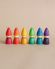 Six colorful, sustainable Grapat Rainbow Tomten figures lined up in a row, each consisting of a cone-shaped top and a rounded base, displayed against a plain, light beige background.