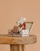 The Maileg Big Sister On the Go Suitcase Set features a small mouse in an orange striped outfit sitting in a floral-patterned suitcase, holding a heart shape. A plain beige background emphasizes this charming scene on a wooden table.