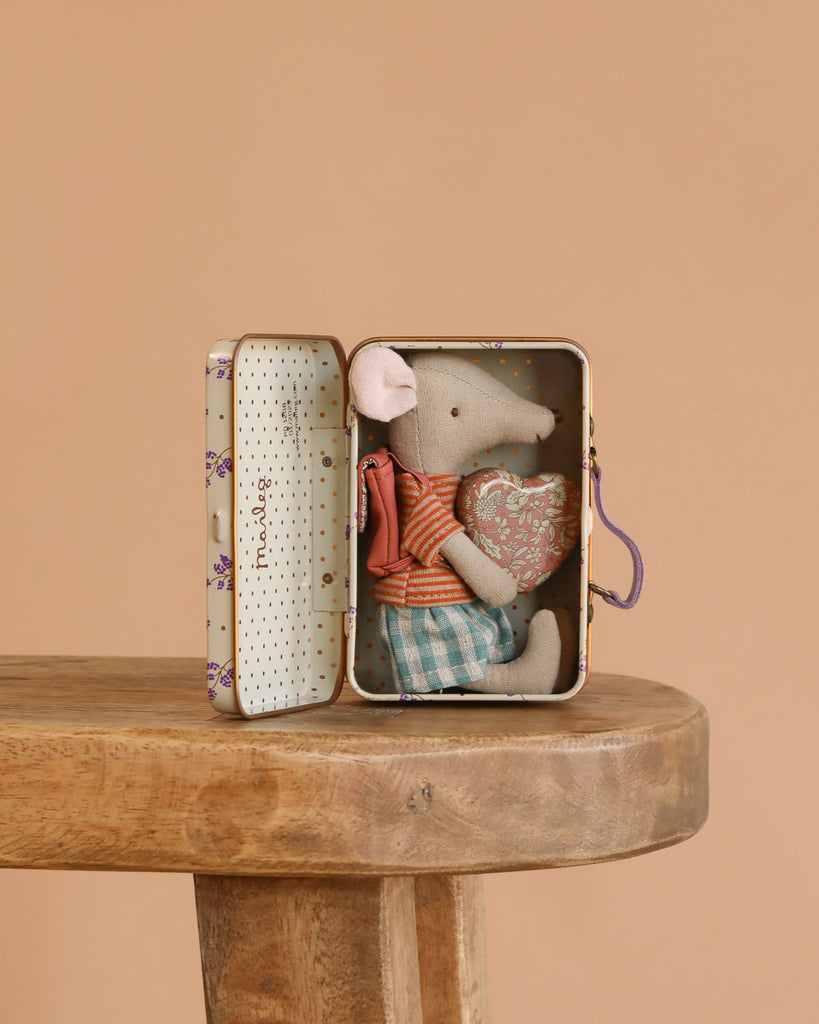 The Maileg Big Sister On the Go Suitcase Set features a small mouse in a striped shirt and plaid shorts, sitting inside an open suitcase on a wooden stool, hugging a heart pillow against a plain beige background.