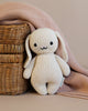 A Cuddle + Kind Baby Bunny toy with long floppy ears sitting next to a wicker basket, partially covered by a soft pink cloth on a beige background.
