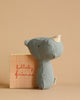 A soft, dusty blue stuffed animal toy resembling a rhino from the Maileg Lullaby Friend Rattles collection stands next to a small orange and beige book with "lullaby friends" written on the cover. The background is a solid light brown.
