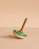 The Mader Bonbon Spinning Top - Green features a charming green and cream wooden design, displayed on its side against a beige background, highlighting the timeless appeal of this classic Austrian toy.