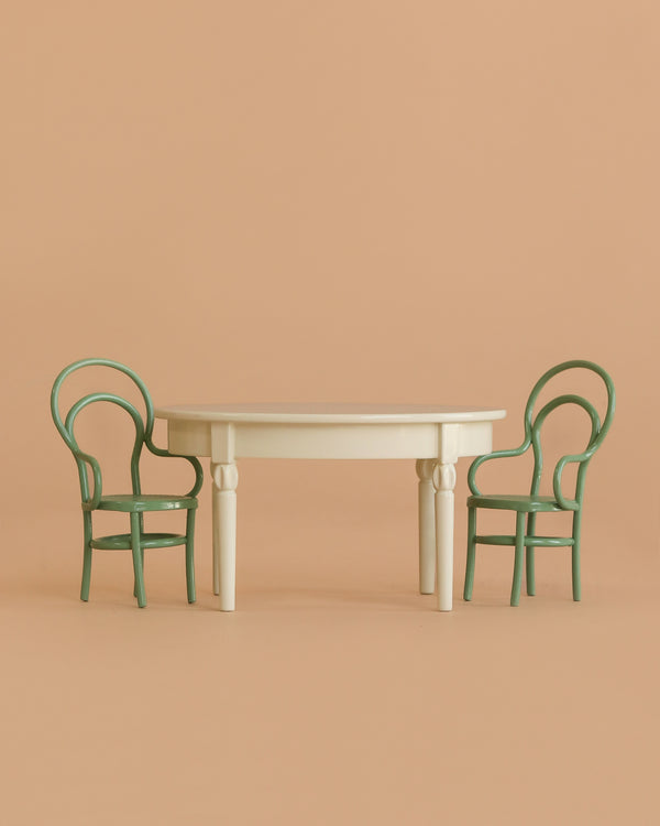 A Maileg Dining Set in cream, sized for mice, includes a vintage table placed between two green round-backed chairs against a plain beige background.