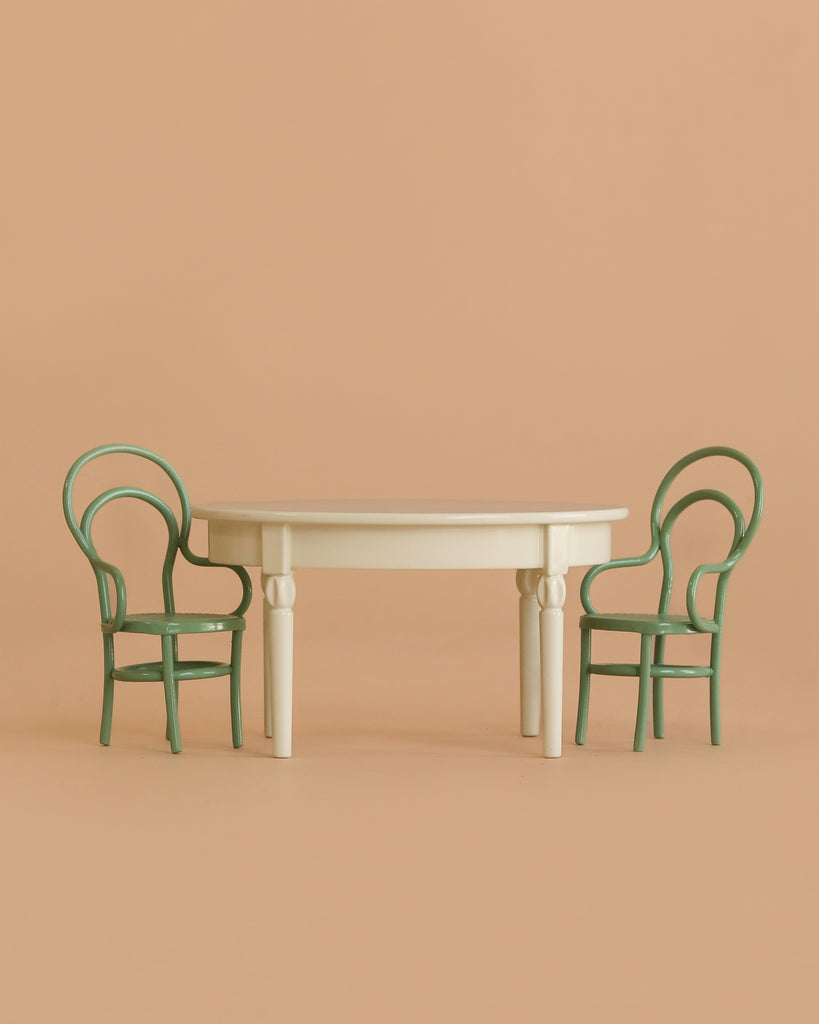 A minimalist scene featuring the Maileg Little Brother and Sister Dining Set, with a cream round vintage table and two green curved-back chairs arranged on either side, set against a solid beige background.