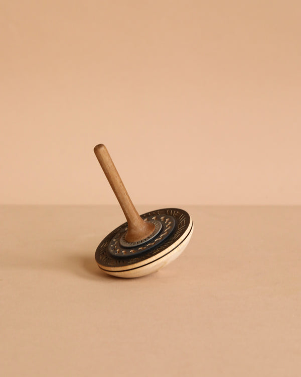 The Mader Bonbon Spinning Top - Black, with its embossed pattern, rests tilted on a beige surface against a plain beige background, capturing the timeless charm of classic Austrian toys.
