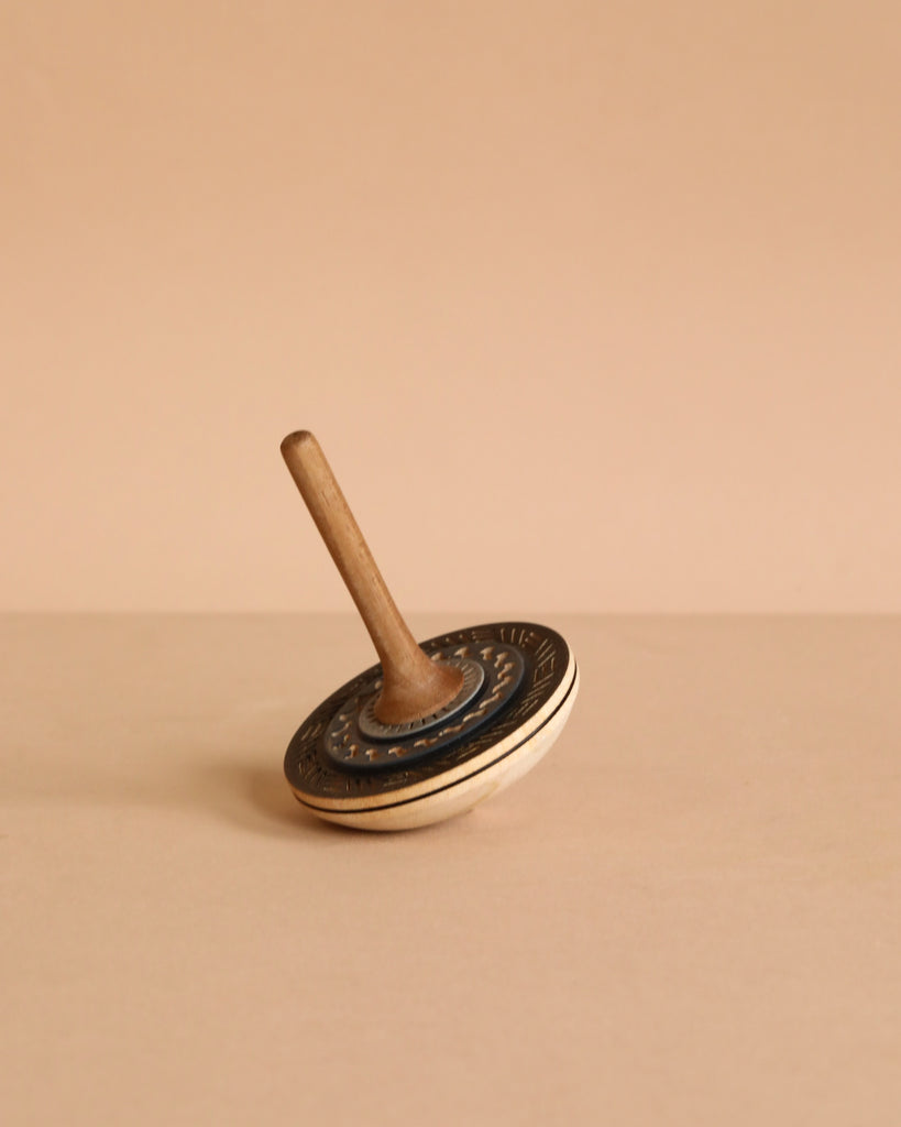 The Mader Bonbon Spinning Top - Black, with its embossed pattern, rests tilted on a beige surface against a plain beige background, capturing the timeless charm of classic Austrian toys.
