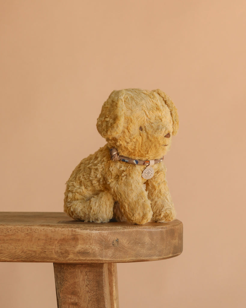 The Maileg Puppy in Dusty Blue Pet Carrier - Soft Sand sits on a wooden bench, its soft fur and patterned collar contrasting with the plain beige background.
