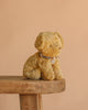 The Maileg Puppy in Coral Pet Carrier - Soft Sand, a plush toy with a decorative collar, sits on a wooden bench against a plain beige background.