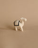 A small, minimalist Handmade Wooden Camel with abstract black patterns stands on a beige background. The figurine, reminiscent of handcrafted wooden animals, has a rectangular patterned cloth depicted on its back and is designed in a simplified, stylized form.
