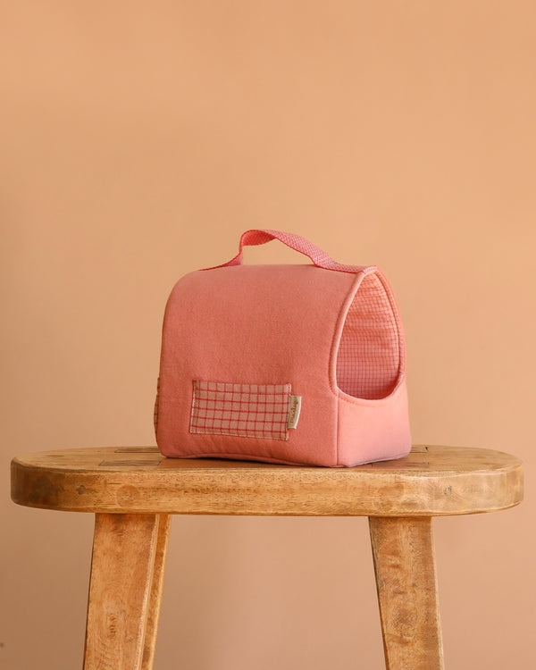 The Maileg Pet Carrier - Coral, a small pink cotton-linen carrier with a grid-patterned pocket and handle, sits on a wooden stool against a plain beige background.