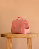 The Maileg Pet Carrier - Coral, a small pink cotton-linen carrier with a grid-patterned pocket and handle, sits on a wooden stool against a plain beige background.
