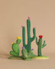 The Bumbu Wooden Cacti Set features a delightful array of clay and wooden cactus sculptures, showcasing different shapes and vibrant colors. One cactus has yellow dots with yellow flowers, another green one adorns small pink birds, a third is speckled with white spots and topped with a red flower, and the last is a short, wide piece crowned with purple flowers.