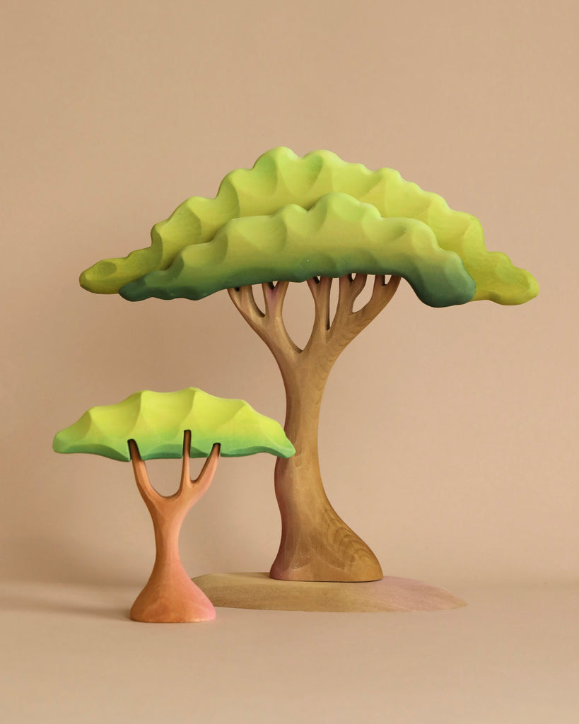 Two stylized wooden trees with green canopies and brown trunks stand on a beige surface—perfect for imaginative play. This handcrafted set pairs beautifully with the Bumbu Handmade Wooden Prehistoric Set.