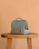 The Maileg Puppy in Dusty Blue Pet Carrier - Dusty Powder, with a handle and patterned front pocket, sits on a wooden stool against a beige backdrop. Its interior includes soft, beige leopard-print fabric—ideal for dog accessories.