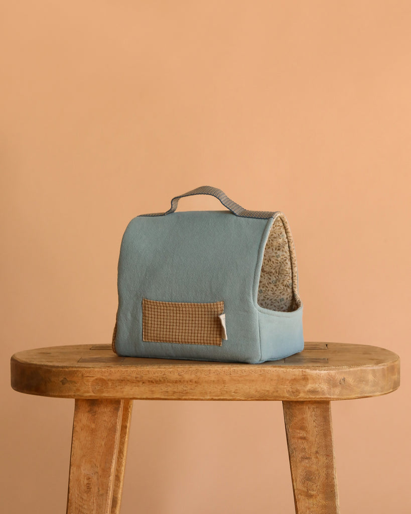 The Maileg Puppy in Dusty Blue Pet Carrier - Dusty Powder, with a handle and patterned front pocket, sits on a wooden stool against a beige backdrop. Its interior includes soft, beige leopard-print fabric—ideal for dog accessories.