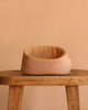 The Maileg Small Sleeping Kitten in Basket—a peach-colored, round pet basket with yellow and white striped lining—rests on a wooden stool against a plain beige background.