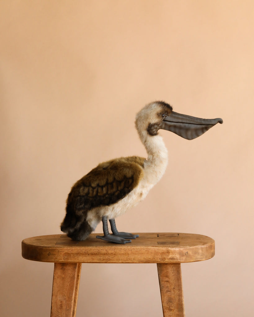 The Pelican Stuffed Animal, a realistic brown and white plush toy with a large beak, sits on a wooden stool against a beige background—ideal for fans of hand-sewn stuffed animals.