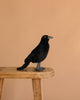 The Crow Bird Stuffed Animal, a black plush toy from HANSA, sits on a light wooden stool against a plain beige background.