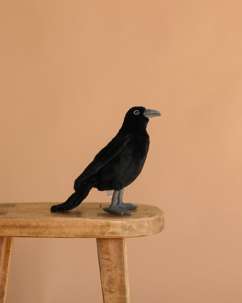 The Crow Bird Stuffed Animal, a black plush toy from HANSA, sits on a light wooden stool against a plain beige background.