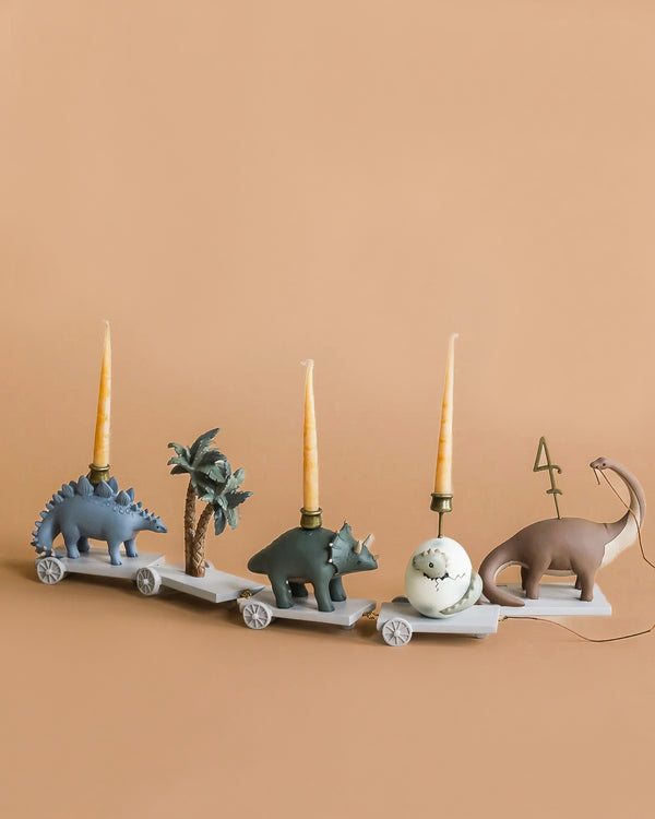 The Dinosaur Birthday Train With Beeswax Candles features four wheeled cars with dinosaurs or a palm tree; two dinosaurs hold yellow candles, one has an ornament, and the last displays an interchangeable number "4"—perfect for dinosaur parties.