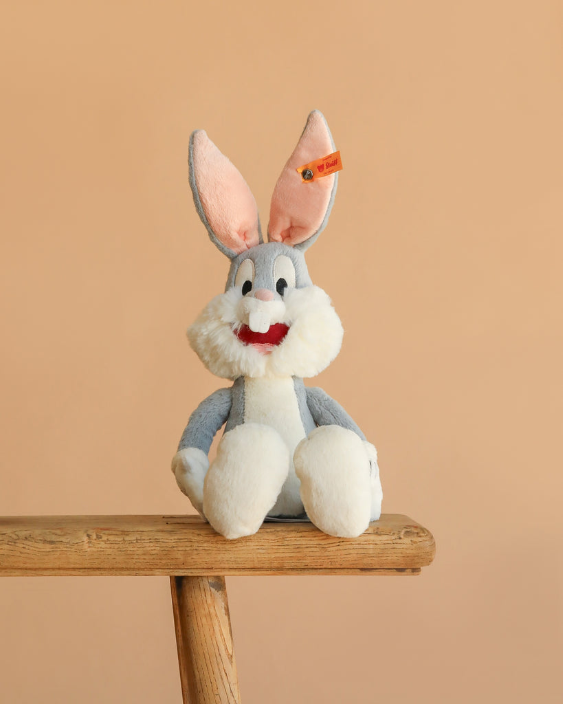 The Steiff Looney Tunes Bugs Bunny plush, featuring gray and white fur, large pink ears, and a red smiling mouth, sits on a wooden stool against a plain beige background.