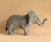 The Ride-On Elephant is a large, artisan hand-sewn plush with realistic features—white tusks, raised trunk, and slightly open mouth—shown standing on a plain light brown background.