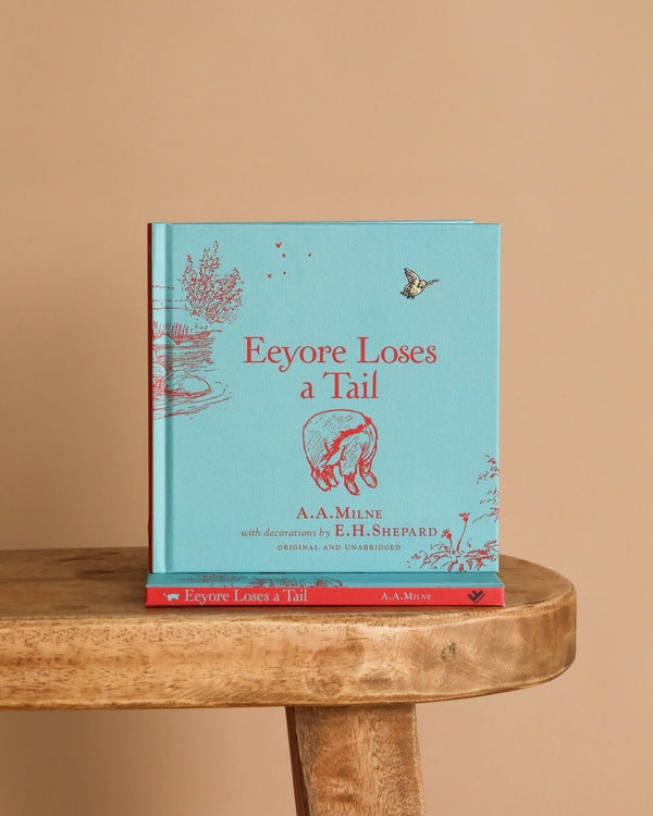 A blue hardcover "Winnie-the-Pooh: Eeyore Loses a Tail" book with red illustrations and text by A.A. Milne stands upright on a wooden stool, with another copy lying flat beneath it against a beige background.