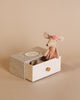 The Maileg Big Sister and Me Set features a doll in a pink dress and headband, sitting inside an open textured box with a small drawer, set against a neutral beige background.
