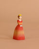 The Bumbu Handmade Wooden Princess features a woman in a yellow crown and red-orange dress with hands folded, ideal for Waldorf-inspired play, shown against a beige background.