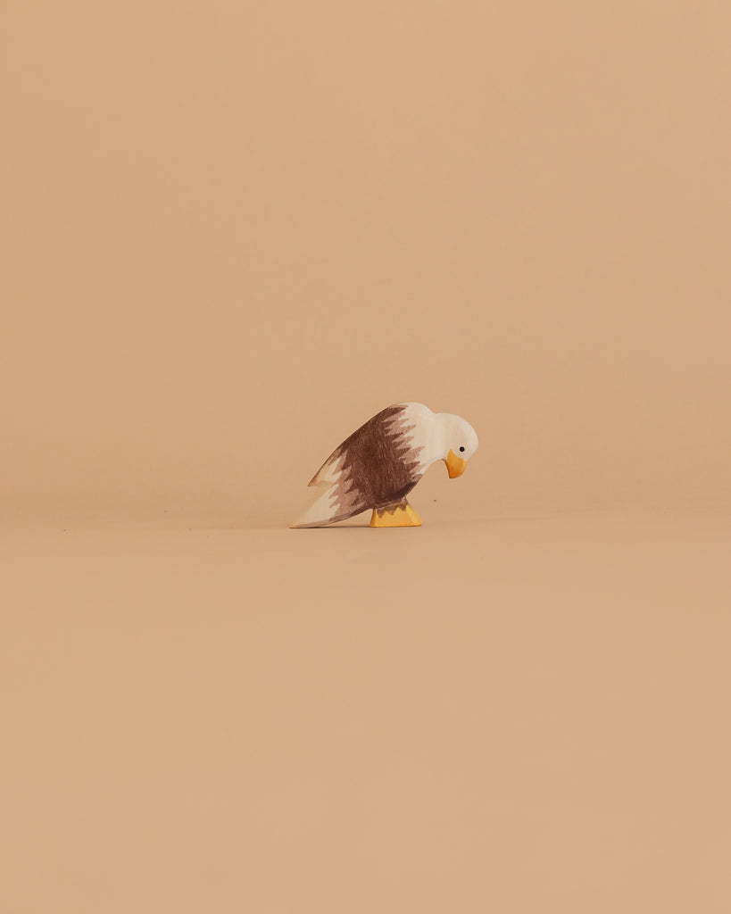 The Ostheimer Bald Eagle Family Set features a small handcrafted wooden figurine with a white head, brown body, and yellow beak, standing on a beige surface against a matching background—perfect for classic toy collections.