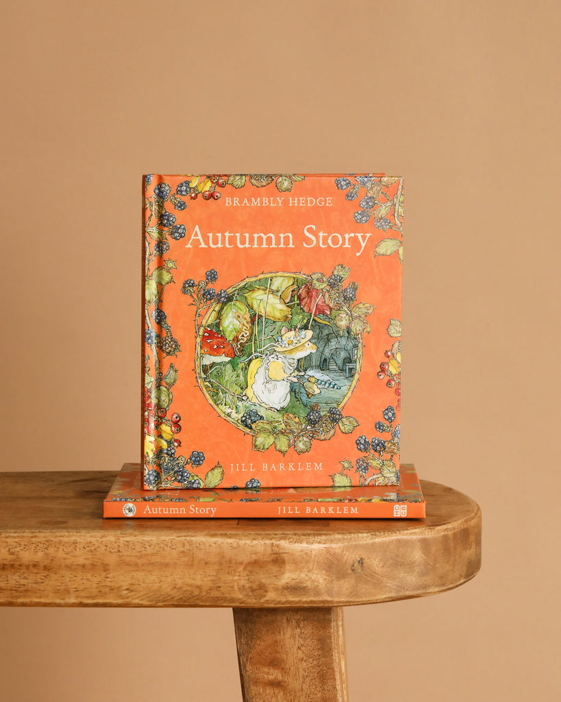 The hardcover "Autumn Story : Jill Barklem" stands upright on a wooden bench with another copy below. Its cover features an illustrated mouse among autumn harvest, berries, and leaves in the magical Brambly Hedge.