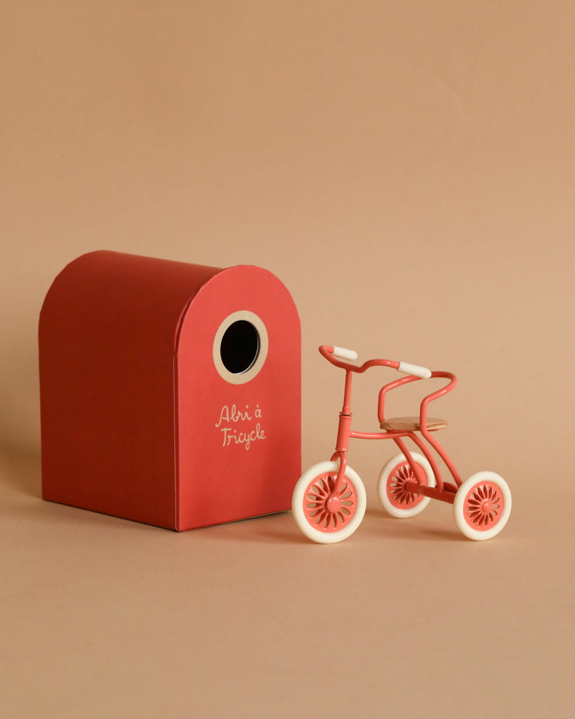 The Maileg Big Sister Tricycle Set features a small red and white tricycle beside a matching arched-roof red box labeled "Abri à Tricycle," all set against a beige background.