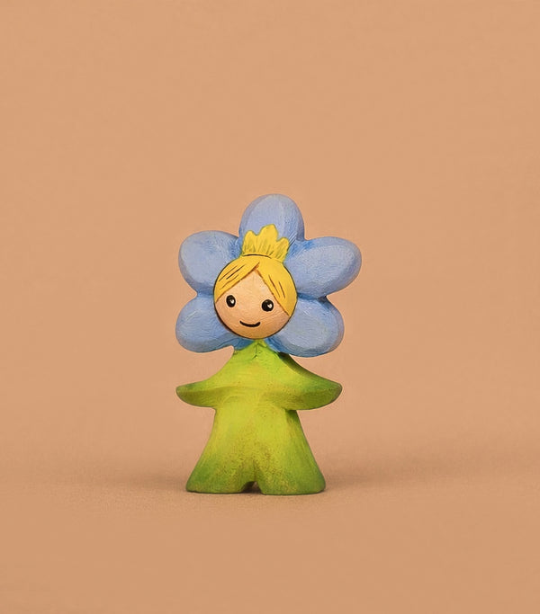The Handmade Wooden Flower Child Forget-Me-Not, with a smiling face, yellow hair, blue flower head, and green leaf outfit, stands against a beige background—ideal for fostering emotional intelligence in Waldorf toy collections.