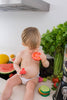 A toddler in a diaper sits on a kitchen counter holding Renato the Tomato Teething Toy, surrounded by fresh fruits and veggies like watermelon, celery, and tomatoes, playfully pretending to eat.