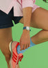 A person in a pink shirt, blue shorts, and a Mini Kyomo Watch - Blue Ocean with Japanese movement bends one knee against a green background.