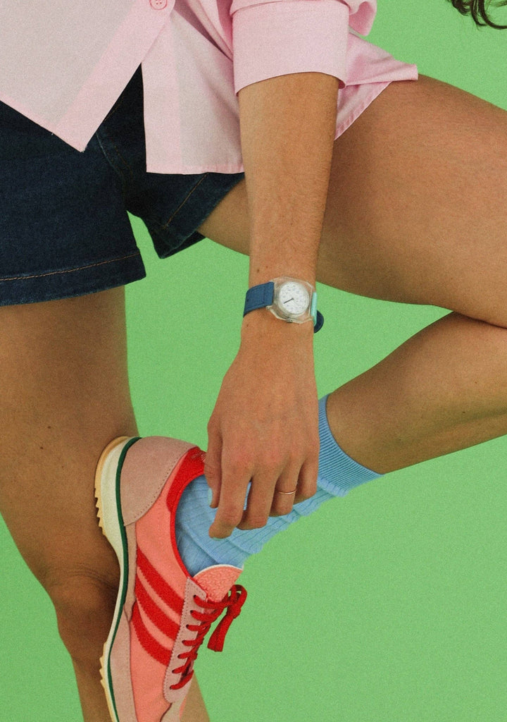 A person in a pink shirt, blue shorts, and a Mini Kyomo Watch - Blue Ocean with Japanese movement bends one knee against a green background.