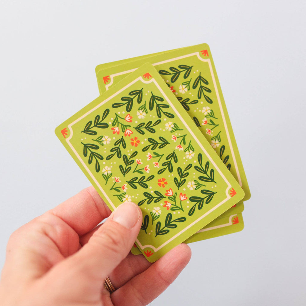 A hand holds two Green Floral Deck of Playing Cards (ships in approximately one week), featuring hand-illustrated leafy and floral motifs on a plain light background—perfect for game enthusiasts.