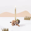 A unique Lion Cake Topper shaped like a lion, crafted from fine porcelain, featuring a lit slender taper candle on its back, positioned against a minimalist background with abstract brown mountains and green cutouts.