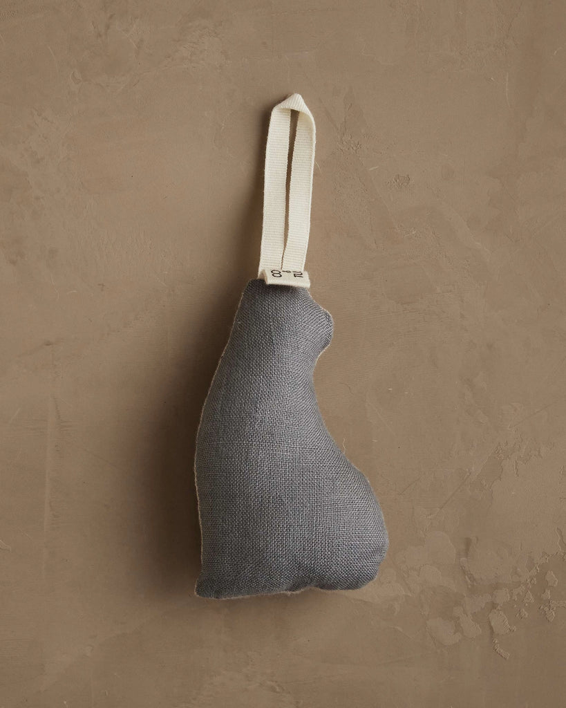 The Coral & Tusk Bear with Tinsel Ornament—a soft, dark gray, irregularly-shaped bear with a white looped strap—hangs against a textured, light brown wall, adding woodland charm to your holiday home decor.