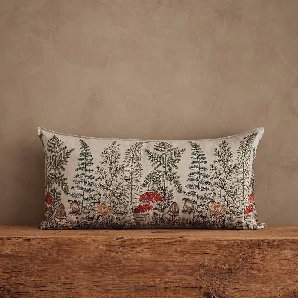 The Coral & Tusk Mushrooms and Ferns Lumbar Pillow is a rectangular embroidered linen accent featuring botanical mushrooms and ferns in earthy tones, displayed on a rustic wooden surface against a beige textured wall.