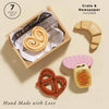 The Bakery & Patisserie Wooden Market Crate includes four pretend pastries—a palmier, croissant, pretzel, and iced bun—displayed in a wooden crate lined with newspaper print. Text: "7 Piece Set," "Crate & Newspaper Included," and "Hand Made with Love.