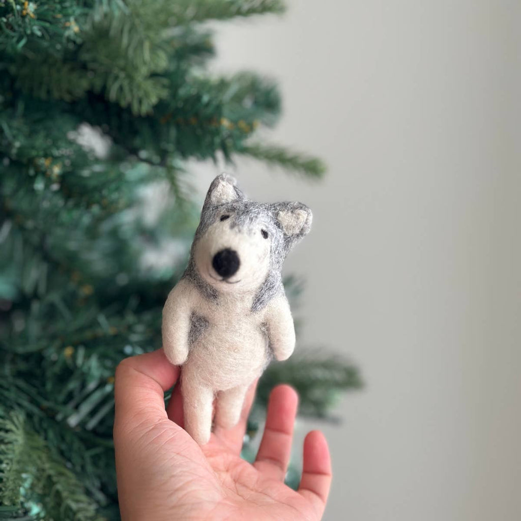 A hand holds a Felt Finger Puppet: Alaskan Husky Dog in front of a green Christmas tree, with blurred branches in the background.