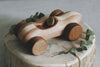 A Handmade Wooden Race Car sits atop a tree stump slice, surrounded by a few green leaves. The toy car has a simple, rustic design with ultra smooth curves and light and dark wood tones, finished with natural linseed oil.